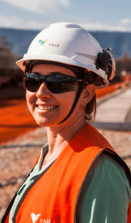 Foto tirada de busto para cima de uma mulher sorrindo em uma área operacional. Ela usa camisa verde clara, colete laranja, óculos de proteção e capacete branco com logotipo da Vale.