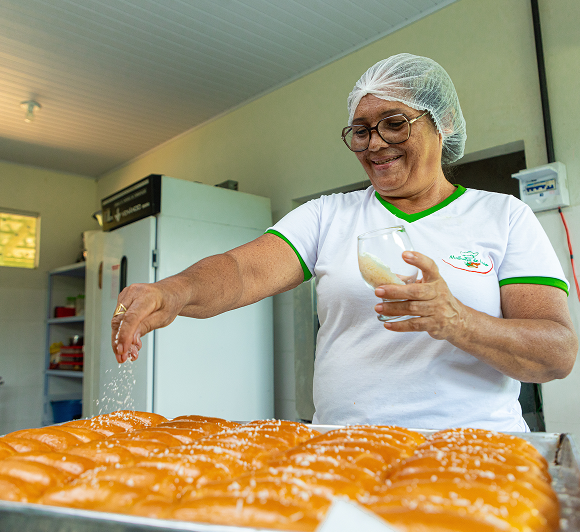 The image shows a person wearing a white shirt with green trim and a hairnet, sprinkling what appears to be powdered sugar or another fine substance onto a tray of baked goods. The person's face is obscured. In the background, there is a refrigerator and some shelves with various items.