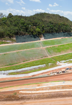 The image shows a landscape with a hillside covered in vegetation and several terraced sections. There are patches of green grass or crops on the terraces, and some areas appear to be under construction or development with visible dirt paths and machinery. The sky is clear with some clouds, and there is a forested area at the top of the hill.