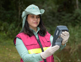 The image shows a person wearing a green hat, a light green long-sleeve shirt, and a pink vest with yellow reflective stripes. The person is holding a handheld device and appears to be outdoors in a natural setting. The person's face is blurred.