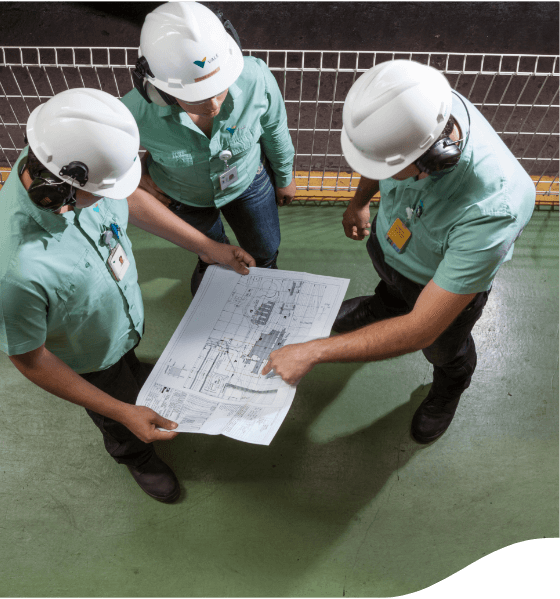 Three individuals wearing safety helmets and green shirts stand on a green floor, looking at and discussing a large blueprint or technical drawing held by one of them. A white fence is visible in the background.