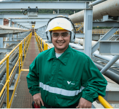 Person on a metal footbridge in an operational area, wearing a white helmet with company logo, safety goggles, ear protection, and a dark green uniform, representing safety practices in energy generation and distribution operations.