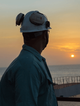A person wearing a hard hat and safety glasses is looking toward the horizon during sunset. They are dressed in a blue work uniform and appear to be standing near a body of water, with industrial structures in the background.