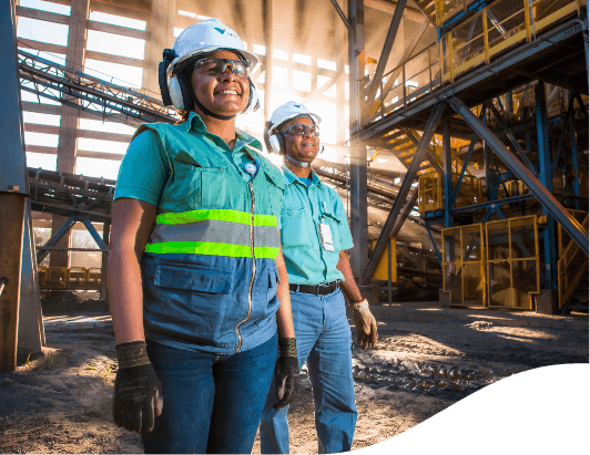 Two workers wearing helmets, gloves, and protective eyewear in an industrial setting lit by sunlight, surrounded by metal structures and conveyor belts.