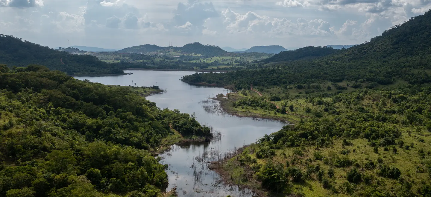 Wide aerial landscape view of a winding river or reservoir surrounded by dense green native forests and rolling hills under an overcast sky.