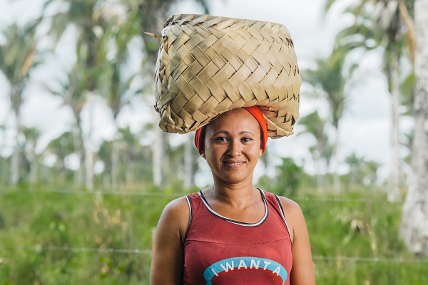 Uma mulher equilibra um cesto de palha na cabeça e sorri para uma fotografia. No fundo, há algumas árvores