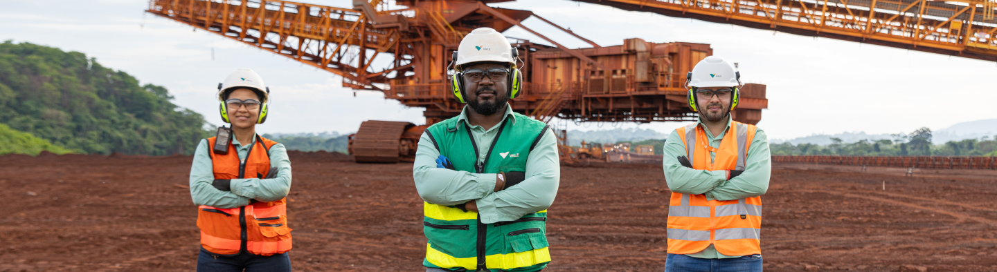 Três profissionais em um ambiente de mineração, vestindo capacetes, óculos de proteção, protetores auriculares e coletes de segurança. Eles estão de braços cruzados em frente a um grande maquinário industrial em um terreno avermelhado, com vegetação ao fundo.