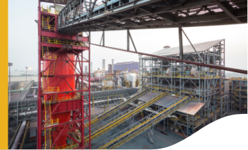 The image shows an industrial facility with multiple interconnected structures, including a prominent red tower on the left side and several conveyor belts and metal frameworks. Various pipes, platforms, and walkways are visible throughout the complex. The background includes additional buildings and industrial equipment.