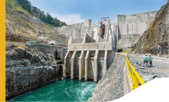 A large hydroelectric dam with water flowing through its spillways. The structure is made of concrete and is situated in a mountainous area. Two workers are visible on the right side of the image, standing near a yellow railing that runs along the edge of the dam. The water below is a deep green color, indicating its depth and flow rate.