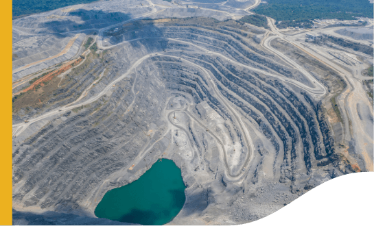 Aerial view of a large open-pit mine with terraced levels and winding roads. There is a turquoise-colored water body at the bottom of the pit. The surrounding area includes more roads and some vegetation in the distance.