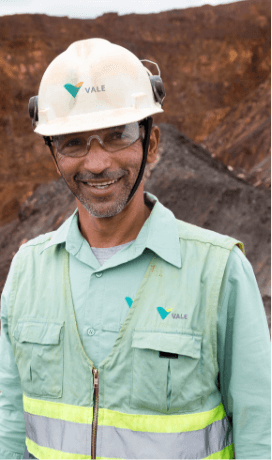 A person wearing a white hard hat with the "VALE" logo and a light green uniform with reflective strips. The background appears to be an outdoor industrial or mining site.
