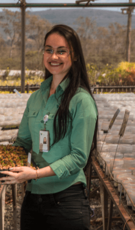A person standing in what appears to be a greenhouse or plant nursery. The person is wearing a green shirt and black pants, with an identification badge clipped to the shirt. The person's face is obscured. In the background, there are rows of plants on tables and some trees visible through the greenhouse structure.