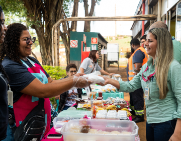The image shows a scene of people at an outdoor market or fair. There are tables set up with various items, including food and drinks. One person is handing an item to another person across the table. The background includes trees and buildings, with other individuals visible in the scene.