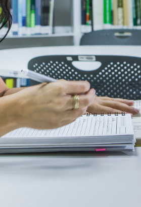 A person is writing in a notebook with a white pen. The notebook is open on a white table, and in the background, there is a black chair with a perforated backrest and several bookshelves.