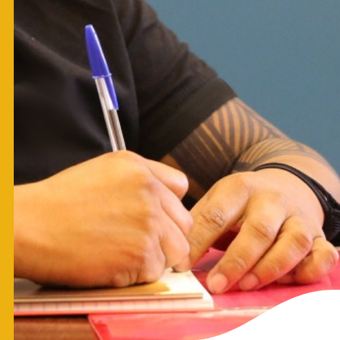 A person is writing in a notebook with a blue pen. Their hands and part of the arm are visible — one hand holds the pen while the other rests on the notebook. The person is wearing a black shirt, and a tattoo is visible on the forearm.