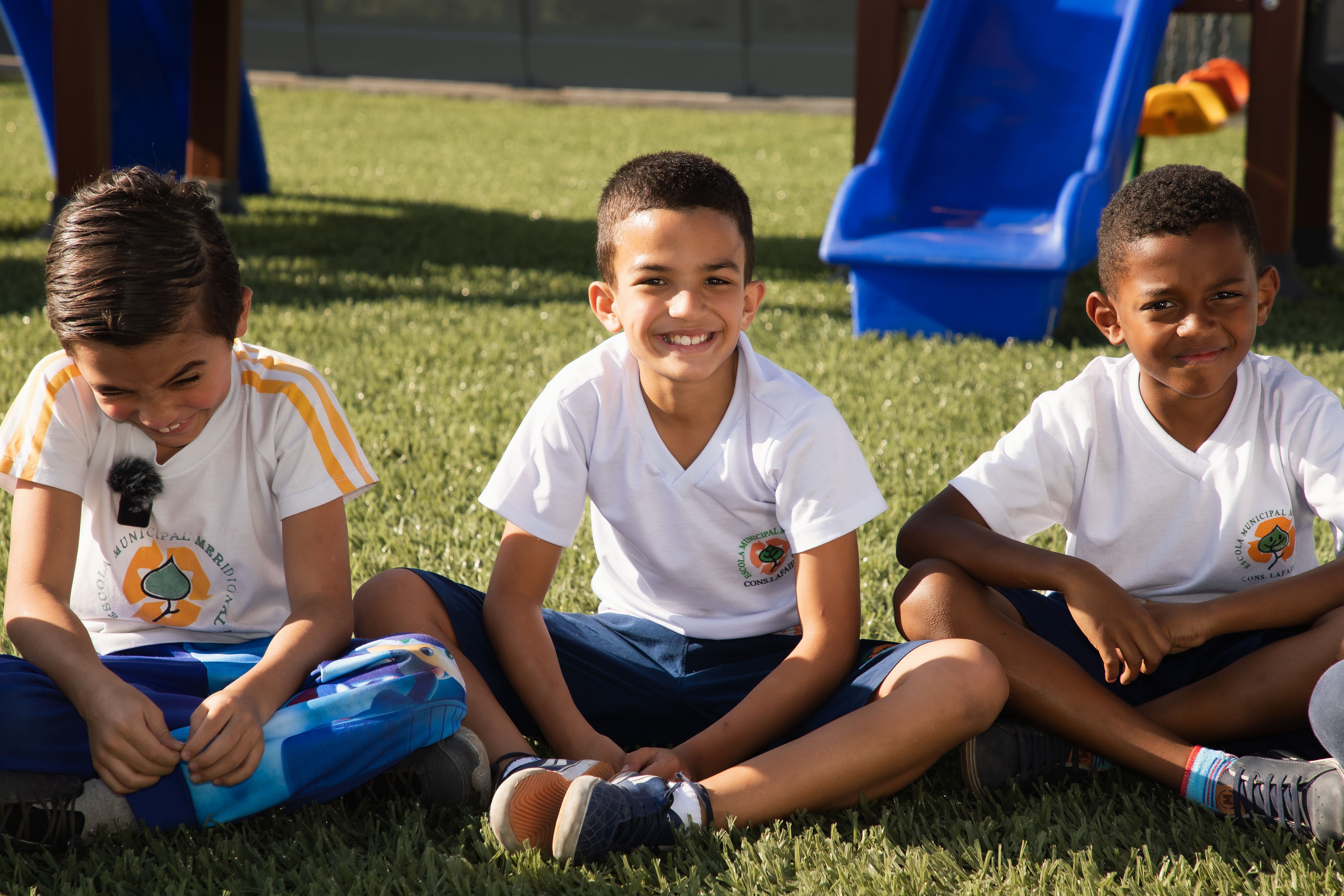 Three children are sitting on the grass, wearing white shirts with a logo and blue pants. In the background, there is playground equipment, including a blue slide.