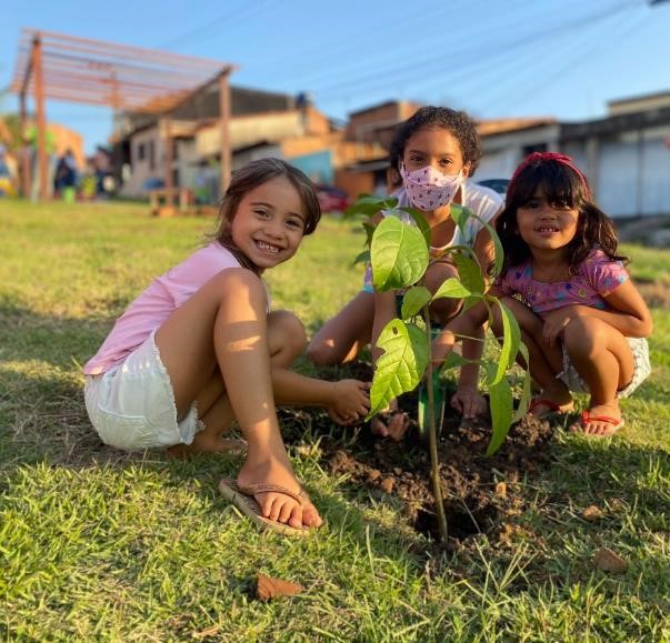 Image of three girls crouched on a lawn, all in front of a tree seedling.