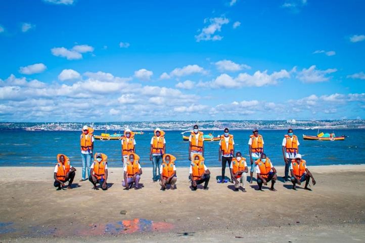Image of several people, seven standing and eight crouched on a beach. All use orange life vests.