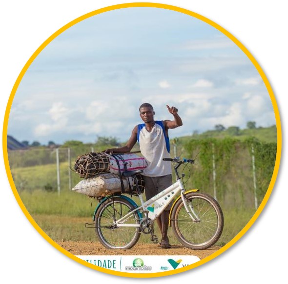 Image of a man, standing, leaning on a bicycle that is loaded with some packages. He gives a thumbs up and the landscape behind is rural.