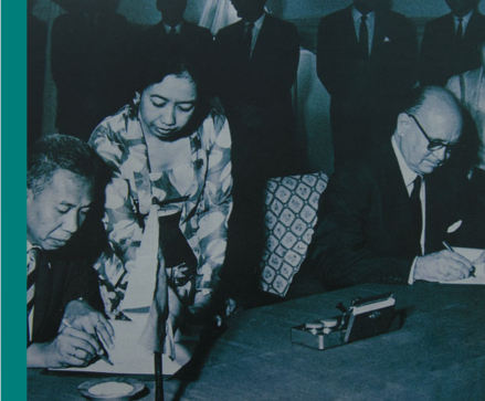 A man in glasses and a suit signs an agreement while a man and a woman in a flowered blouse check other documents.