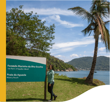 Photo of a woman next to a sign with a coconut tree and one in the background. The woman is wearing Vale uniform, a green long-sleeved button-down shirt, dark pants and sneakers.