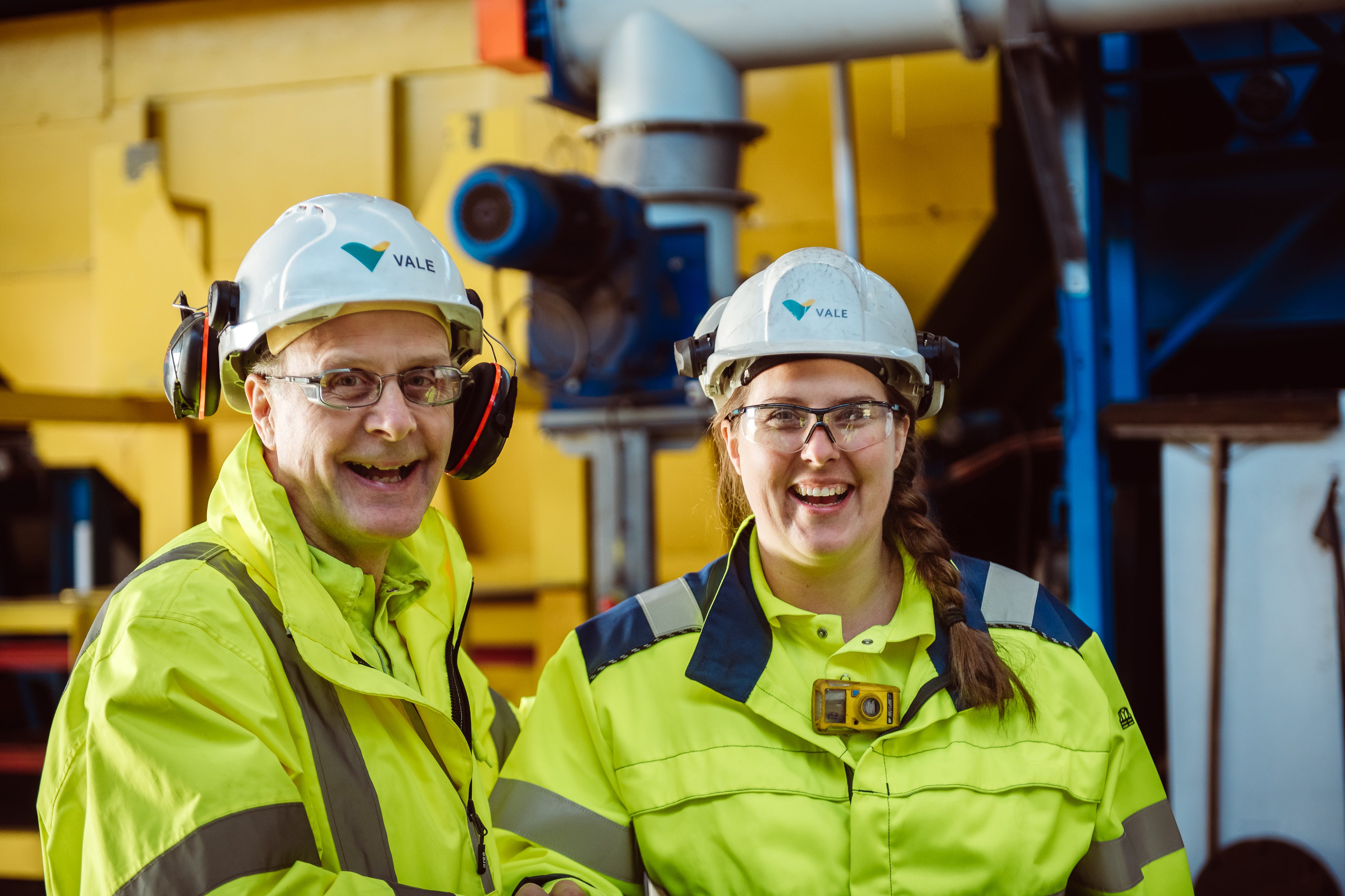 Two employees smiling to the picture. There's an eldry men and a women, both of then wears protections equipaments