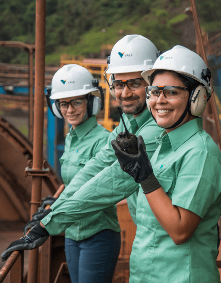 Três empregados Vale, duas mulheres e um homem, estão em uma área externa de operação. Todos usam luvas, capacetes e óculos de proteção e uma das mulheres segura um radiotransmissor.