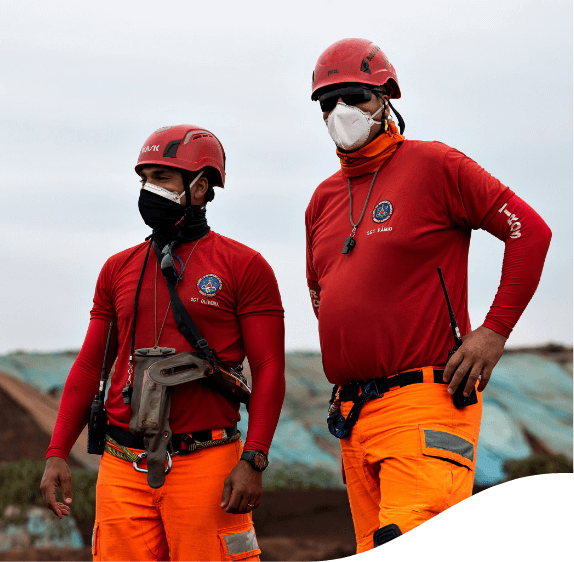 Foto de dois homens bombeiros. Eles estão usando camisa de manga comprida vermelha, calça laranja fluorescente, capacete, máscara e rádio preso na cintura.