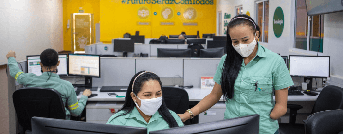 Duas empregadas Vale conversam em um escritório. Uma está sentada e a outra de pé. Ambas usam máscara.