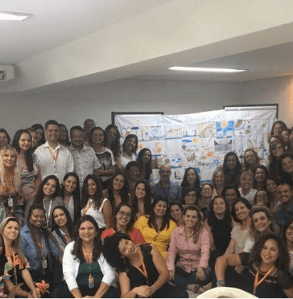 Grupo grande de pessoas, homens e mulheres, posando para foto em frente a um painel numa sala de aula.