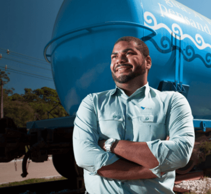 Homem negro sorrindo e olhando para frente. Ele está usando camisa verde clara com logotipo da Vale e atrás dele há uma estrutura de metal em azul e branco.