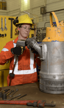 A Vale employee is wearing protective glasses, helmet and gloves. She's operating a piece of equipment in a large iron works