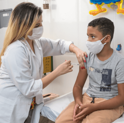 Foto de uma profissional da saúde segurando o braço de uma criança com uma das mãos e aplicando uma injeção com a outra. Ambas usam máscara facial.