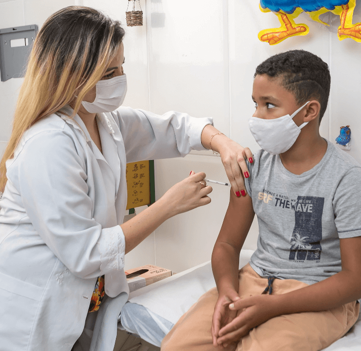 Foto de uma profissional da saúde segurando o braço de uma criança com uma das mãos e aplicando uma injeção com a outra. Ambas usam máscara facial.