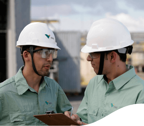 Dois homens conversando em uma área de operação. Um deles está segurando uma prancheta. Os dois estão usando camisa verde clara de botões da Vale, óculos de proteção e capacete.