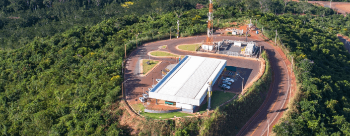 Em um local cercado por árvores, há um grande galpão e uma torre de energia, além de uma estrada.