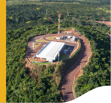 Em um local cercado por árvores, há um grande galpão e uma torre de energia, além de uma estrada.