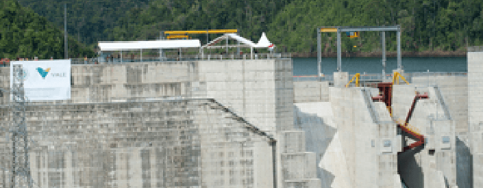 In the image, it is possible to see a kind of concrete wall, with some machinery. On top there is the presence of water, and in the background a mountainous area with dense vegetation.