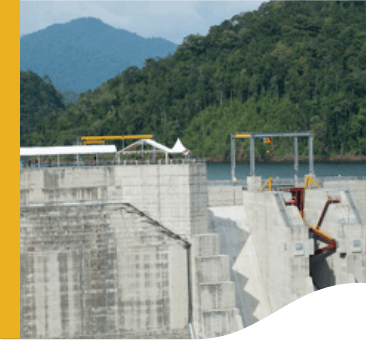 In the image, it is possible to see a kind of concrete wall, with some machinery. On top there is the presence of water, and in the background a mountainous area with dense vegetation.