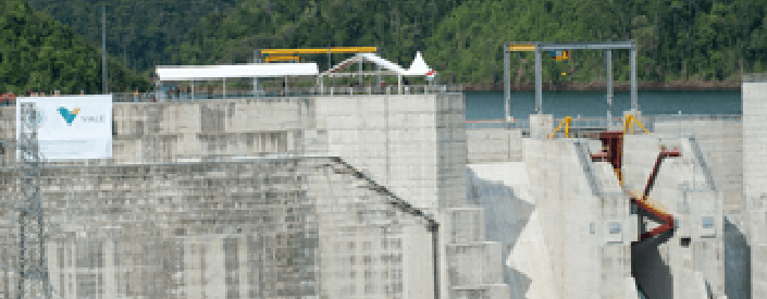 Na imagem é possível ver uma espécie de murro de concreto, com alguns maquinários. Em cima há a presença de água e, ao fundo uma área montanhosa com vegetação densa.