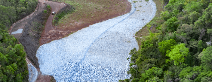 Nas laterais da imagem é possível uma área de vegetação enquanto, no meio, há um caminho coberto por pedras.