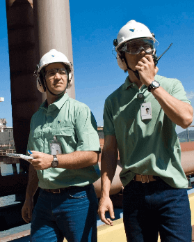 Dois homens lado a lado em um espaço de operações. Os dois usam calças jeans, camisas verdes, óculos de proteção, protetores de ouvido e um capacete branco com logotipo da Vale. Um deles segura um rádio comunicador em uma das mãos.