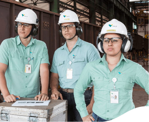 Two men and a woman stand side by side looking at what is in front of them. They all wear light green uniforms, ear protectors, goggles, and white helmets with the Vale logo on them.