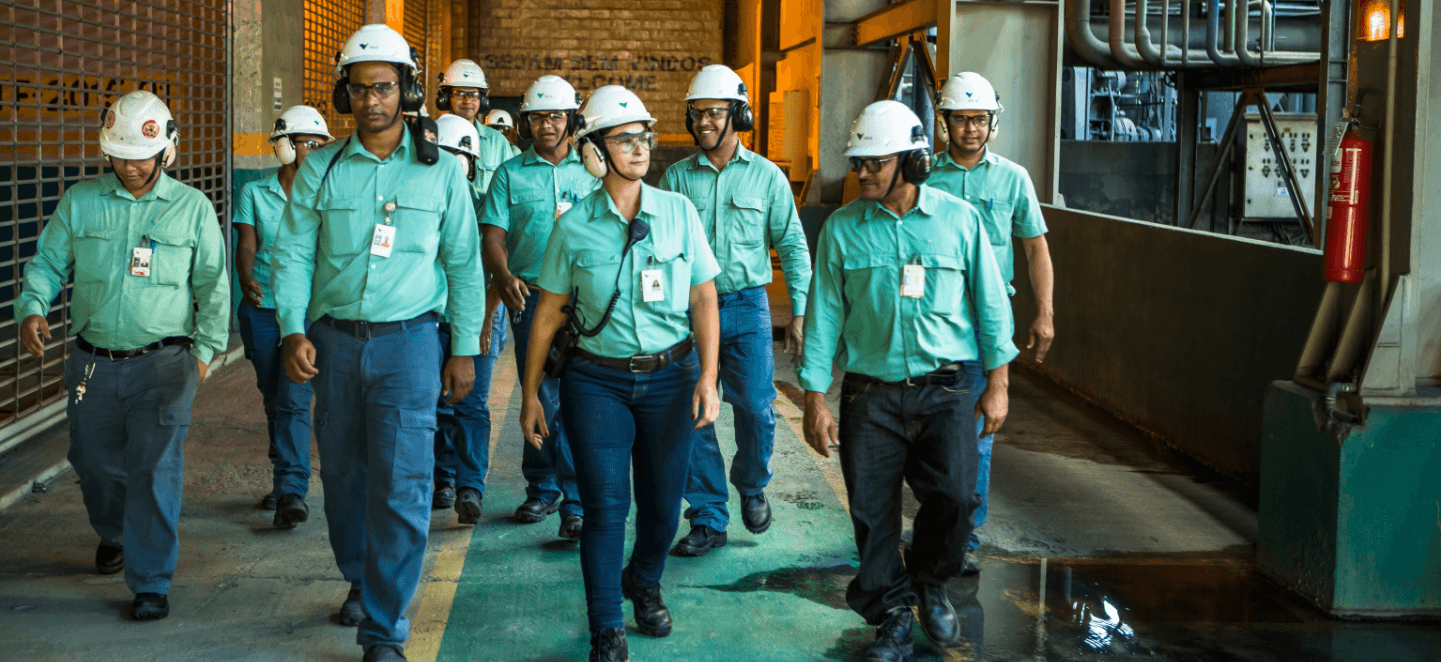 Several Vale employees walking side by side in an operational space. All are wearing, green uniforms, badges, goggles, ear muffs and white helmets with the company logo.