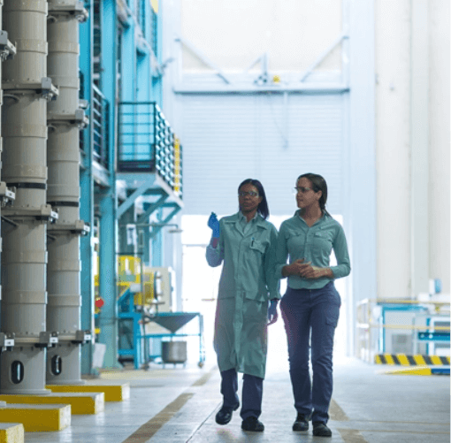 Two Vale employees walk through a large space, which has large metal structures. One of them wears a lab coat and protective gloves, and the other wears jeans pants and the uniform shirt.