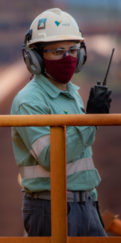 Foto da barriga até a cabeça de uma mulher segurando um rádio em uma operação. A mulher está usando uniforme Vale, capacete, óculos, proteção nos ouvidos e máscara no rosto.