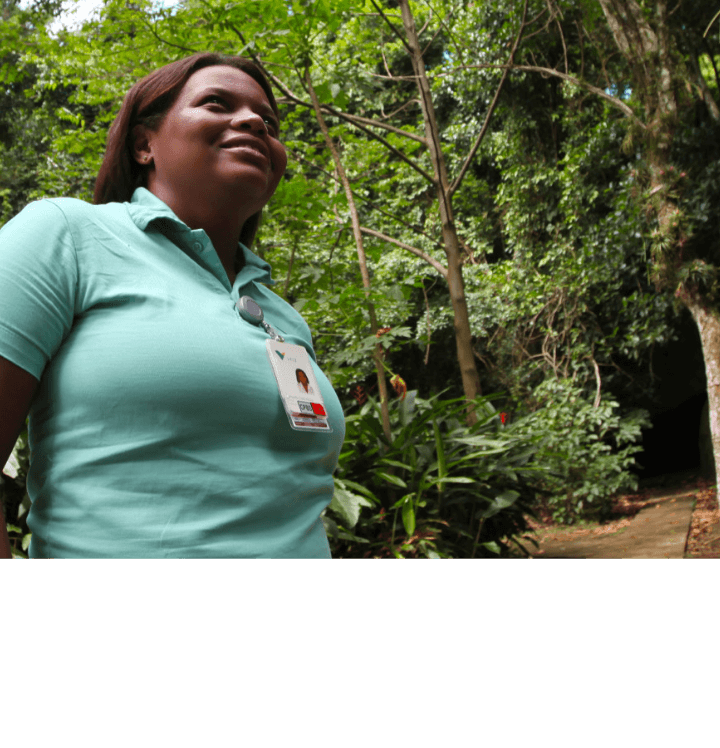 Mulher negra sorrindo em meio a uma área arborizada. Ela usa camiseta verde clara e crachá.