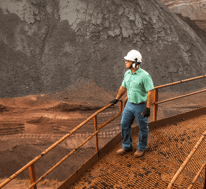 Photo of a man on metal walkway in an operation area with ore and earth in the background. The man is leaning against the grid and is wearing a green button-down shirt, jeans, gloves, ear protection, a helmet and goggles.