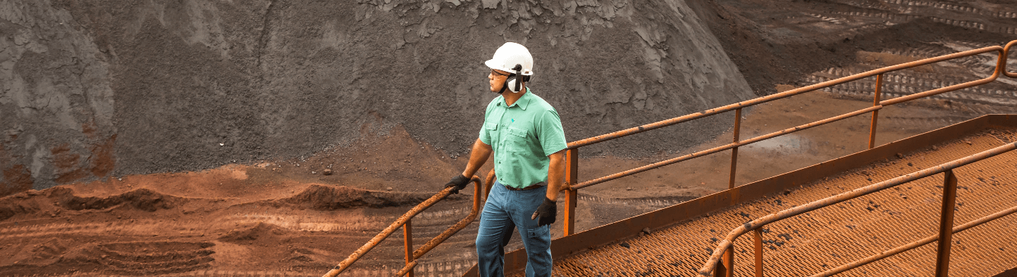Photo of a man on metal walkway in an operation area with ore and earth in the background. The man is leaning against the grid and is wearing a green button-down shirt, jeans, gloves, ear protection, a helmet and goggles.