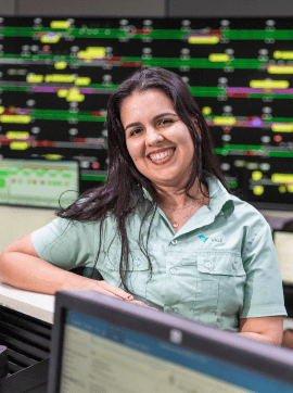 Photo of a woman in an office with multiple monitors in the background. She's leaning against a counter and is wearing a green button-down shirt with Vale's logo, long straight hair and is smiling.